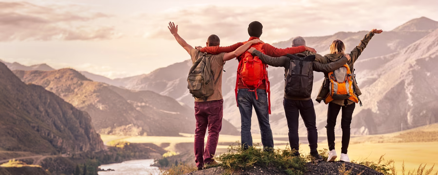 Four friends with backpacks standing on a mountain ledge overlooking a scenic valley, celebrating their outdoor hiking adventure with arms around each other.