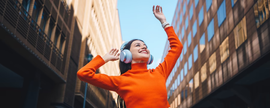 Woman in bright orange sweater smiling and dancing outdoors with headphones on, representing confidence, empowerment, and self-expression.