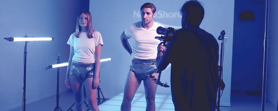 Two models, a woman and a man, stand barefoot in a brightly lit studio with blue lighting, wearing adult diapers. A photographer films them from the foreground while studio lights and equipment surround the set.