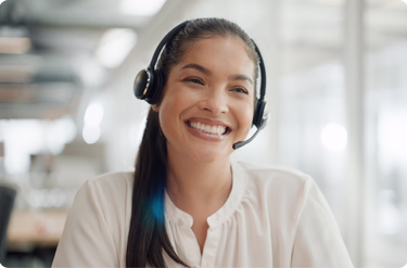 A smiling woman wearing a headset with a microphone looks engaged while working in a bright office setting.